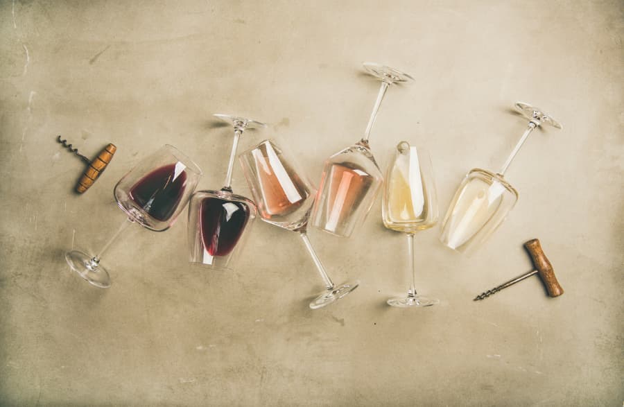 Six wine glasses with red, pink, and light yellow liquid in them with corkscrews on the side on a beige background.