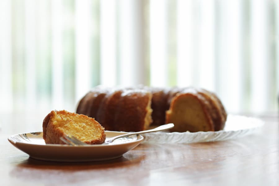 Slice of rum cake on plate with fork with rest of rum cake in background