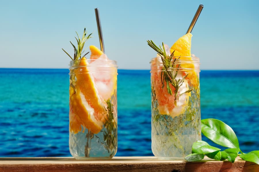 Two glasses of hard seltzer mixed with tropical fruits sitting in a beach setting.